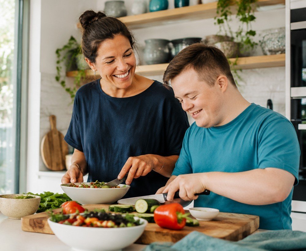 A support worker and a young person laughing together while preparing a meal in a bright home kitchen